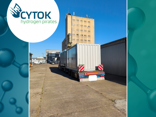 Ein hellgrauer Technikcontainer steht auf einem Tieflader auf dem Gelände eines Industrie- und Forschungsstandorts. Im Hintergrund ist ein mehrstöckiges Gebäude unter blauem Himmel zu sehen. Das Bild ist im Corporate Design von CYTOK gestaltet und zeigt links oben das CYTOK-Logo mit dem Schriftzug „hydrogen pirates“. Die Aufnahme dokumentiert die Anlieferung eines Containers für eine zukünftige modulare Power-to-Gas-Anlage.